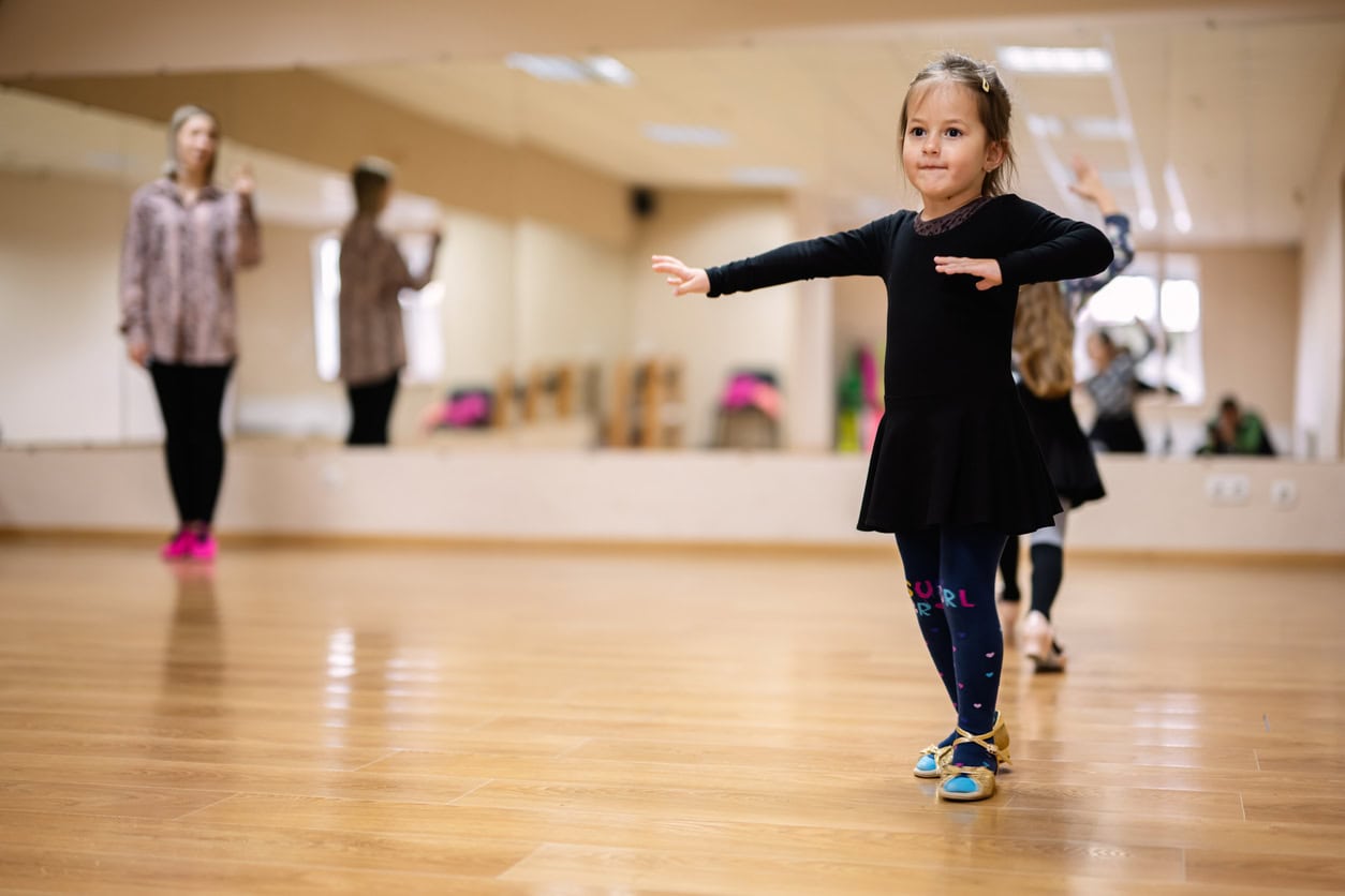Little girl in black dance attire practicing ballet poses in a dance studio with mirrors, guided by her instructor.