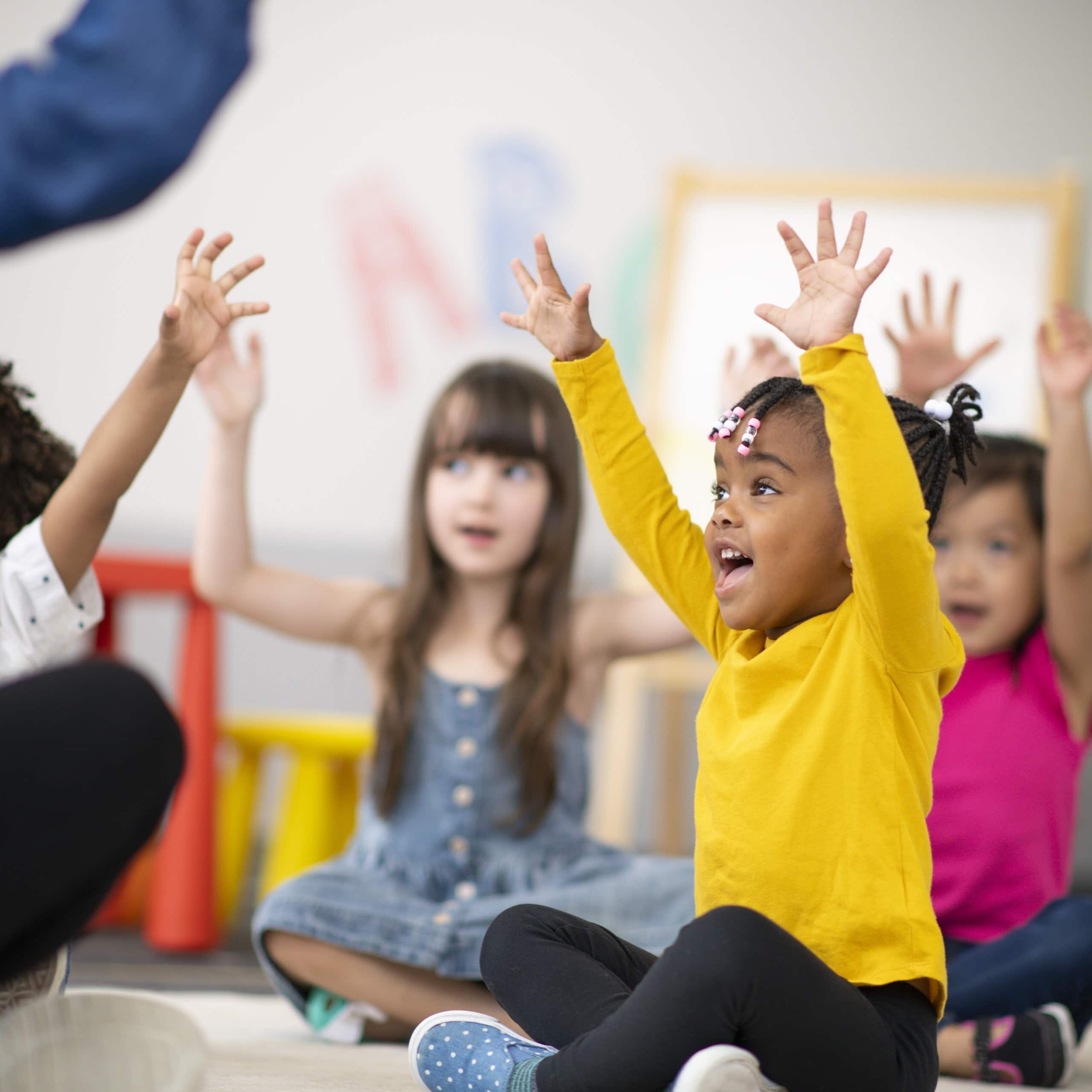 A multi-ethnic group of preschool students is sitting with their legs crossed on the floor in their classroom. The mixed-race female teacher is sitting on the floor facing the children. The happy kids are smiling and following the teacher's instructions. They have their arms raised in the air.